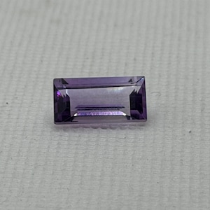 May include: A rectangular, faceted amethyst gemstone. The gem is a deep purple color with a rectangular shape and cut facets. The gemstone is set against a white background.