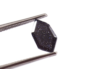 May include: A dark blue gemstone with a faceted surface and sparkling inclusions, held by metal tweezers. The gemstone has an elongated, geometric shape with multiple flat sides. The background is plain white, accentuating the gem's colour and texture.