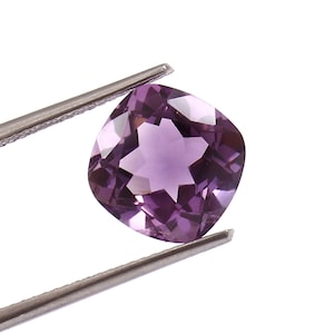May include: A cushion-cut amethyst gemstone held by tweezers. The gemstone is a vibrant purple color with a star-like pattern in the center. The tweezers are silver and the background is white.