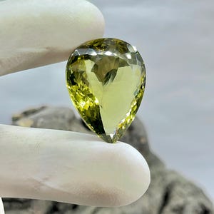 Puede incluir: Una piedra preciosa verde en forma de pera con un corte facetado. La piedra preciosa se sostiene en una mano enguantada.