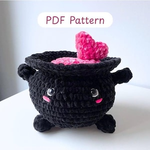 Puede incluir: Un caldero negro de ganchillo con un corazón rosa y líquido rosa en el interior. El caldero tiene una cara sonriente con mejillas rosadas y está etiquetado como "PDF Pattern". Este artículo hecho a mano es perfecto para Halloween.