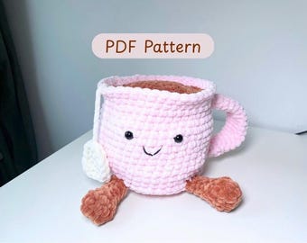 Könnte beinhalten: Eine handgefertigte, lächelnde, rosa gehäkelte Teetasse mit braunem Tee. Sie hat einen Henkel, einen weißen Teebeutel und braune Füße. Der Text "PDF Pattern" steht auf einem hellbraunen Banner.
