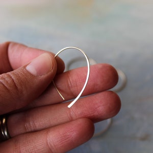 Puede incluir: Un par de pendientes de aro de plata, mostrados en una vista de cerca. Los pendientes son finos y delicados, con un diseño simple y minimalista.