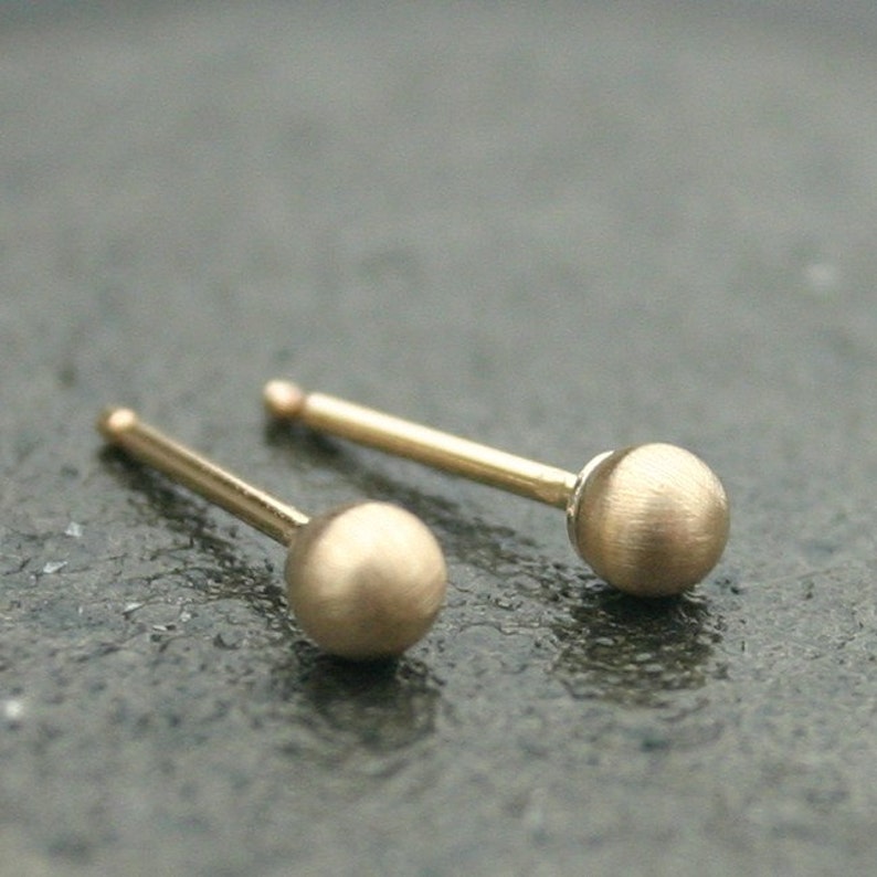May include: A pair of small, gold-colored ball stud earrings. Each earring features a small, round ball at the end of a thin post. The earrings are set against a dark, textured surface.