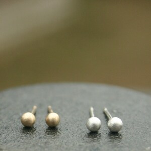 May include: A close-up of two pairs of small stud earrings. One pair is gold-colored, and the other is silver-colored. The earrings have a simple ball design and are set against a dark, textured surface.