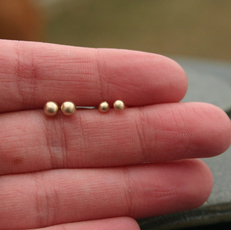 May include: Four pairs of gold-colored ball stud earrings in varying sizes. The earrings are displayed on a person's fingers, showcasing the different sizes of the jewelry. The earrings are small and simple, with a matte finish.