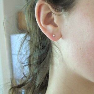 May include: A close-up shot of a person's ear with a small, round, silver stud earring. The ear is positioned on the left side of the frame, with the person's hair partially visible. The earring is a simple, minimalist design.
