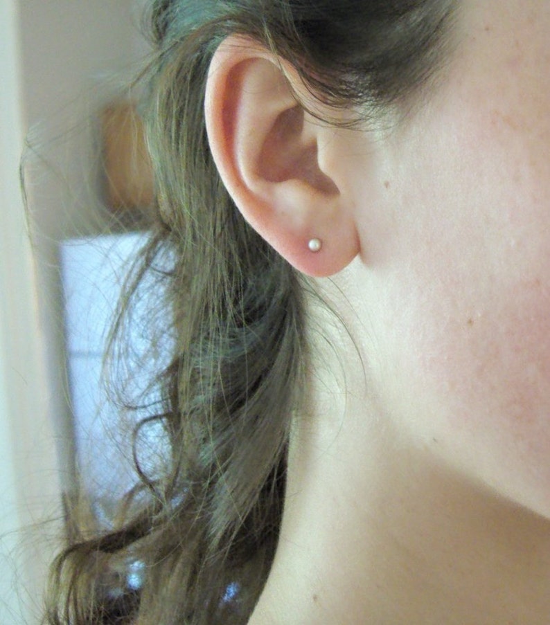 May include: A close-up shot of a person's ear with a small, round, silver stud earring. The ear is positioned on the left side of the frame, with the person's hair partially visible. The earring is a simple, minimalist design.
