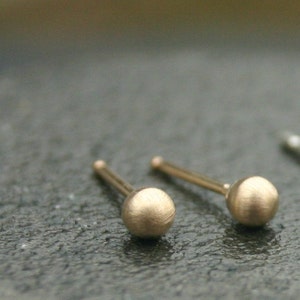 May include: Close-up of two gold-colored stud earrings. Each earring features a small, textured sphere at the end of a thin post. The earrings are set against a dark, textured surface.