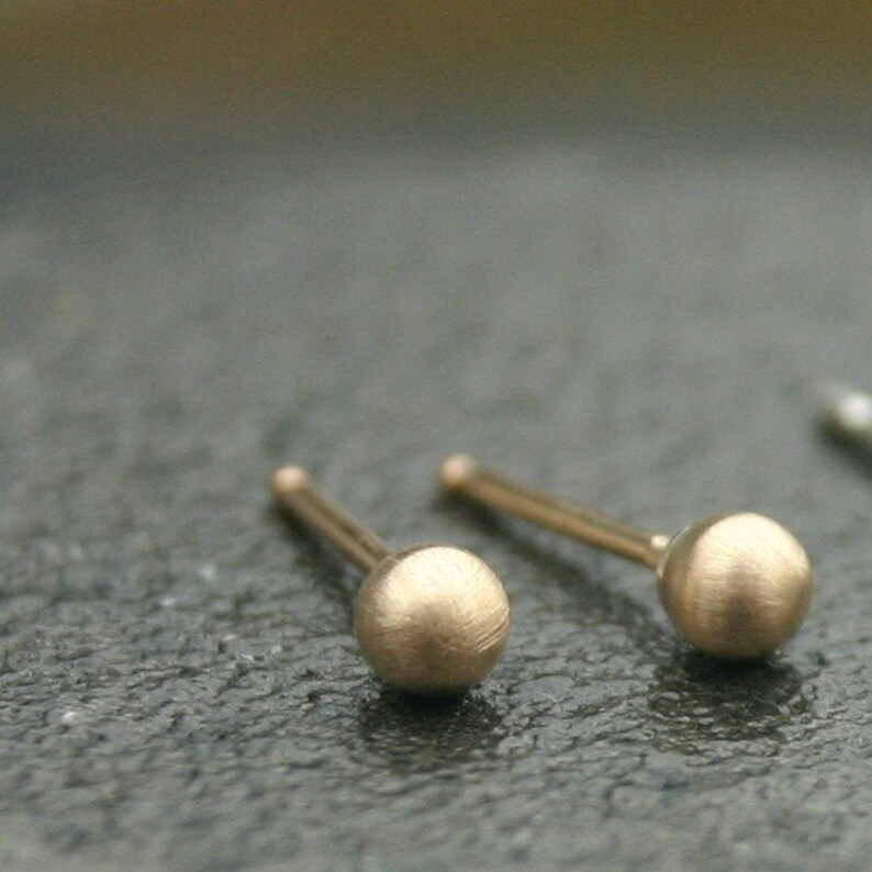 May include: Close-up of two gold-colored stud earrings. Each earring features a small, textured sphere at the end of a thin post. The earrings are set against a dark, textured surface.