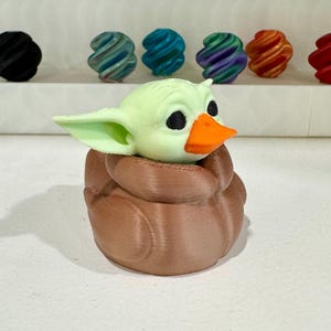Puede incluir: Figura impresa en 3D de un personaje con orejas grandes, ojos negros y un pico naranja, metido en una forma marr&oacute;n, texturizada y envuelta. La figura es de color verde claro. Otros objetos coloridos y arremolinados est&aacute;n en el fondo.