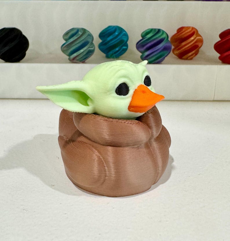 Puede incluir: Figura impresa en 3D de un personaje con orejas grandes, ojos negros y un pico naranja, metido en una forma marr&oacute;n, texturizada y envuelta. La figura es de color verde claro. Otros objetos coloridos y arremolinados est&aacute;n en el fondo.
