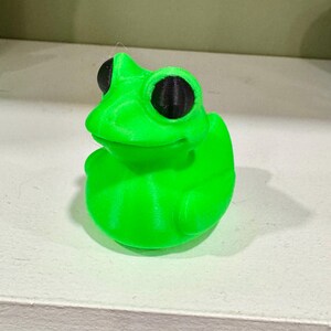 May include: A bright green frog figurine with large black eyes. The frog is sitting upright, with its front legs tucked in. The figurine is made of a smooth, solid material and has a playful, cartoonish appearance.