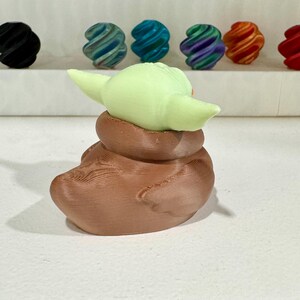 Puede incluir: Una figura impresa en 3D de un personaje con orejas grandes y una t&uacute;nica marr&oacute;n. La figura es de color verde claro y marr&oacute;n. Varias esferas coloridas y arremolinadas est&aacute;n en el fondo, incluyendo negro, verde azulado, azul, morado, naranja y rojo.