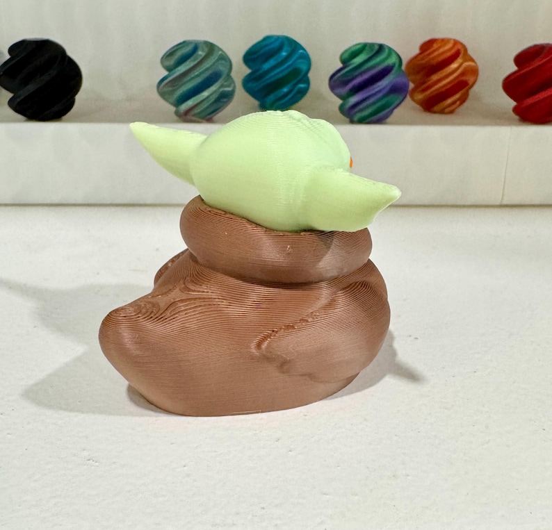 Puede incluir: Una figura impresa en 3D de un personaje con orejas grandes y una t&uacute;nica marr&oacute;n. La figura es de color verde claro y marr&oacute;n. Varias esferas coloridas y arremolinadas est&aacute;n en el fondo, incluyendo negro, verde azulado, azul, morado, naranja y rojo.
