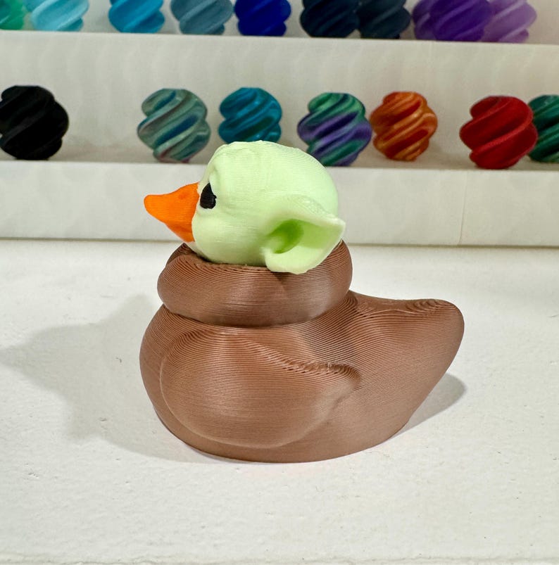 Puede incluir: Una figura de pato marr&oacute;n con cabeza verde claro y pico naranja. El pato tiene una superficie texturizada. En el fondo hay varios objetos decorativos coloridos y arremolinados.