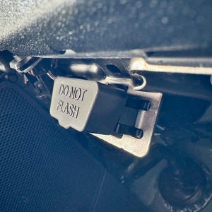 Può includere: Un componente automobilistico nero e argento con la scritta "DO NOT FLASH" stampata sopra. Il componente è fissato all'area del cruscotto di un veicolo, con cablaggio e hardware di montaggio visibili.