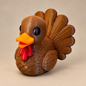 May include: A 3D printed turkey figurine featuring a brown body with textured feather patterns, a bright orange beak, large black eyes, and a vibrant red wattle. The turkey has a fanned tail and a rounded, compact shape.