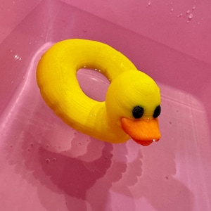 May include: A yellow rubber ducky-shaped pool float with a ring body and a duck head. The duck has black eyes and an orange beak. The float is in a pink tub filled with water.