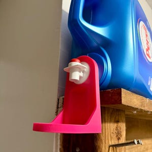 Puede incluir: Un dispensador de detergente para ropa de color rosa brillante con una boquilla blanca y una tapa roja está montado en un estante de madera. Una botella grande de detergente azul está colocada encima del dispensador. El dispensador está diseñado para contener y dispensar detergente líquido.