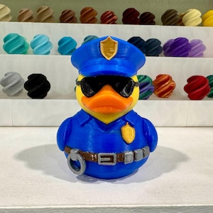 Puede incluir: Un pato de goma amarillo con uniforme de policía azul, gorra y gafas de sol. El pato tiene una placa y un cinturón con esposas. El fondo presenta estantes con varios objetos decorativos coloridos.