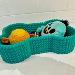 May include: A teal-colored, rope-textured pet bed holds two plush toys. One toy is a yellow and orange ball, and the other is a blue creature with a black mohawk and a cross on its head. The bed is shaped like a figure eight.