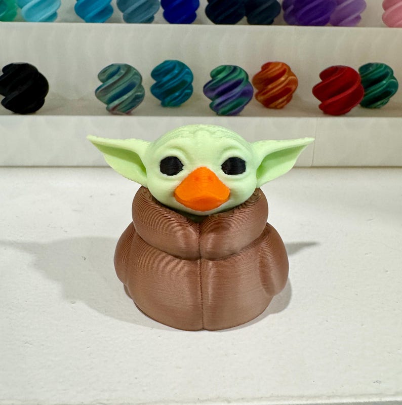 Puede incluir: Una figura verde claro de un personaje con orejas grandes y ojos negros, que lleva una capa marr&oacute;n y un pico naranja. La figura es un h&iacute;brido de un personaje popular y un pato. Varias cuentas coloridas y arremolinadas est&aacute;n en el fondo.