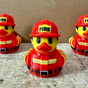 May include: Three rubber duck toys dressed as firefighters. Each duck is wearing a red firefighter helmet with the word "FIRE" on it, and a red uniform with yellow stripes and a black belt.