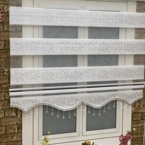 May include: White window blinds with a decorative scalloped bottom edge and crystal-like beads hanging from the bottom. The blinds have a white background with a silver floral pattern.
