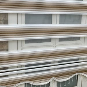 以下が含まれることがあります： ベージュと白のストライプの窓ブラインドに、ビーズの装飾的なトリムが付いています。ブラインドは部分的に上げられており、白い窓枠が見えています。