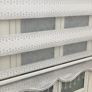 Könnte beinhalten: Weiße Fensterjalousien mit einem zarten Blumenmuster und einer gewellten Volant mit Kristallperlen.