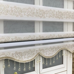 Puede incluir: Persianas blancas para ventanas con un patrón floral dorado y una cenefa de cuentas. La cenefa tiene un borde festoneado y cuelga en una franja de cuentas transparentes.
