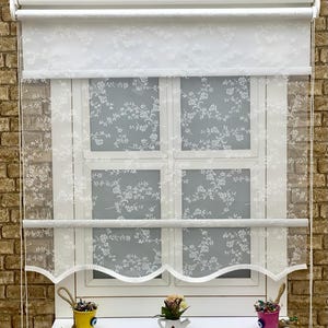 Op de afbeelding: Witte, doorschijnende raamrolluiken met een bloemenpatroon. De rolluiken zijn gedeeltelijk opgerold, waardoor een wit raamkozijn met vier ruiten zichtbaar wordt. De rolluiken hebben een gegolfde onderrand.