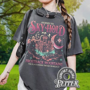 Puede incluir: Camiseta gris oscuro con un diseño gráfico que presenta un castillo, una luna y estrellas, con el texto "JOURNEY TO SKY HOLD" y "FROSTBACK MOUNTAINS". La camiseta tiene un aspecto vintage y la marca "PEPPER" es visible.