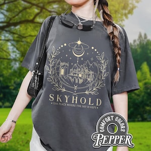May include: Dark grey t-shirt with a gold graphic design of a castle, moon, stars, and the word "SKYHOLD". The text "THE PLACE WHERE THE SKY IS KEPT" is below. The shirt has a Comfort Colors Pepper label.