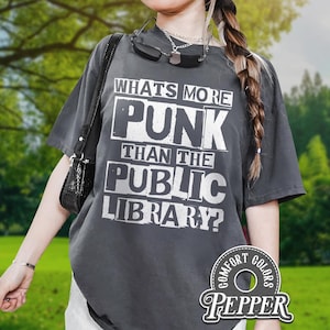 May include: Dark gray t-shirt with the text "WHAT'S MORE PUNK THAN THE PUBLIC LIBRARY?" in white block letters. The shirt has a relaxed fit and is part of the Comfort Colors collection.