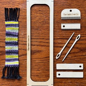 May include: A wooden weaving loom kit with a woven bookmark. The kit includes a large rectangular loom, a small comb, a shuttle, and two needles. The woven bookmark has purple, yellow, and white stripes with black fringe. The text "The Yarn Over llc" is printed on the loom.