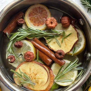 May include: A stainless steel pot filled with water containing sliced yellow lemons, dried apple slices, brown cinnamon sticks, small dried pumpkins, and sprigs of green rosemary. The ingredients are submerged in the liquid for a stovetop simmer.