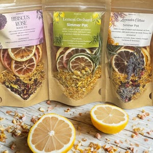 May include: Three simmer pot sachets with dried fruit and herbs, labeled "Hibiscus Rose", "Lemon Orchard", and "Lavender Citrus". Two lemon halves are in the foreground. The sachets are in clear bags.