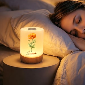 May include: A cylindrical bedside lamp with a warm, glowing white shade featuring a printed orange marigold flower and the name Anna in cursive script. The lamp has a light-colored wooden base and a circular silver-toned button on the top surface.