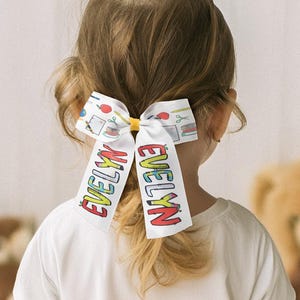 May include: White hair bow with the name "EVELYN" in colourful letters. The bow features a pattern of school supplies and is tied with a yellow band. The bow is worn in a ponytail.