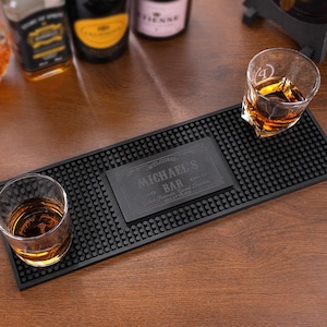 Könnte beinhalten: Eine schwarze Gummi-Bar-Matte mit einer personalisierten Plakette mit der Aufschrift "MICHAEL'S BAR". Zwei Gläser mit bernsteinfarbener Flüssigkeit stehen auf der Matte. Die Matte liegt auf einer Holzoberfläche, im Hintergrund Flaschen.
