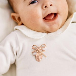 Op de afbeelding: Een religieuze broche van roségoud met een strik, kruis en medaillon is vastgemaakt aan een witte romper. Het delicate accessoire is waarschijnlijk voor een baby. De baby lacht met open mond.