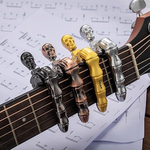 Op de afbeelding: Akoestische gitaar met vijf metalen capo's in de vorm van een schedel, in zwart, brons, goud en zilver. Elke capo is op de gitaarsnaren geklemd, met namen gegraveerd op de zijkanten. Muzieknoten op de achtergrond.