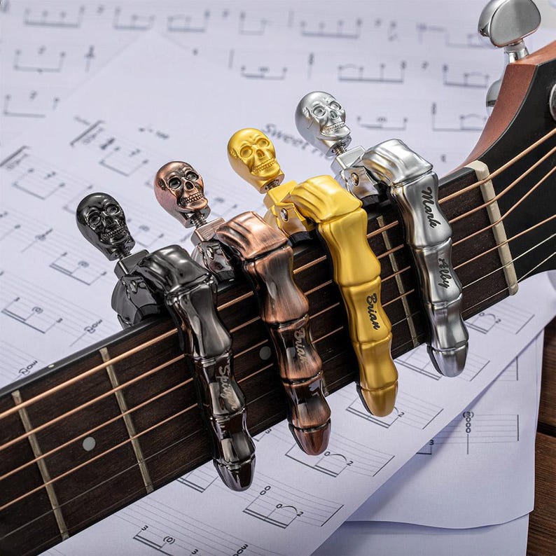 Op de afbeelding: Akoestische gitaar met vijf metalen capo's in de vorm van een schedel, in zwart, brons, goud en zilver. Elke capo is op de gitaarsnaren geklemd, met namen gegraveerd op de zijkanten. Muzieknoten op de achtergrond.