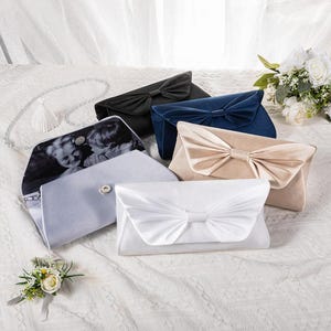 May include: An assortment of elegant clutch purses in various colors, including black, navy blue, beige, and white. Each clutch features a decorative bow on the front. One clutch displays a black and white photo inside.