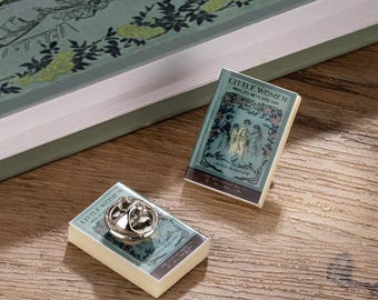 May include: A Little Women book pin and a miniature book with a teal cover and floral design. The pin has a silver-coloured backing. The book cover features illustrations of women and the title "Little Women" by Louisa May Alcott.