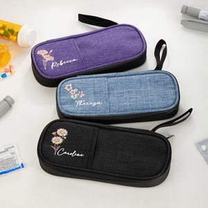 May include: Three personalized pencil cases in purple, blue, and black. Each case has a floral design and a name embroidered on it: Rebecca, Theresa, and Caroline. The cases have a black zipper and a black handle.