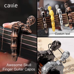 Op de afbeelding: Gitaarkapokken met schedelontwerpen in brons, zilver, goud en zwart. De kapokken zijn bevestigd aan een gitaarhals. De tekst "Awesome Skull Finger Guitar Capos" is zichtbaar.