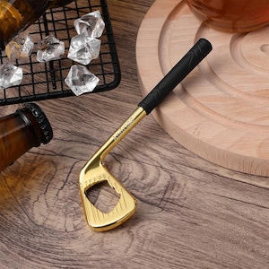 Puede incluir: Un abridor de botellas dorado con forma de palo de golf, con un mango negro. El abridor está sobre una superficie de madera, con cubitos de hielo y una botella en el fondo. El abridor de palo de golf tiene un diseño único.